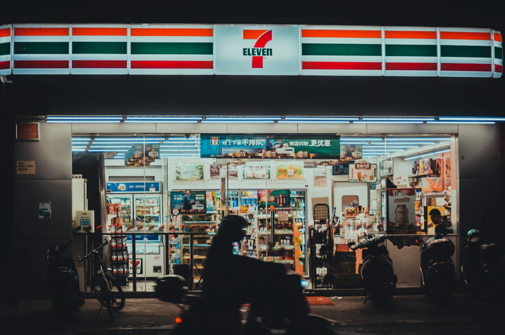 A vibrant 7-Eleven store front lit up during nighttime with a silhouette of a motorbike rider passing by.