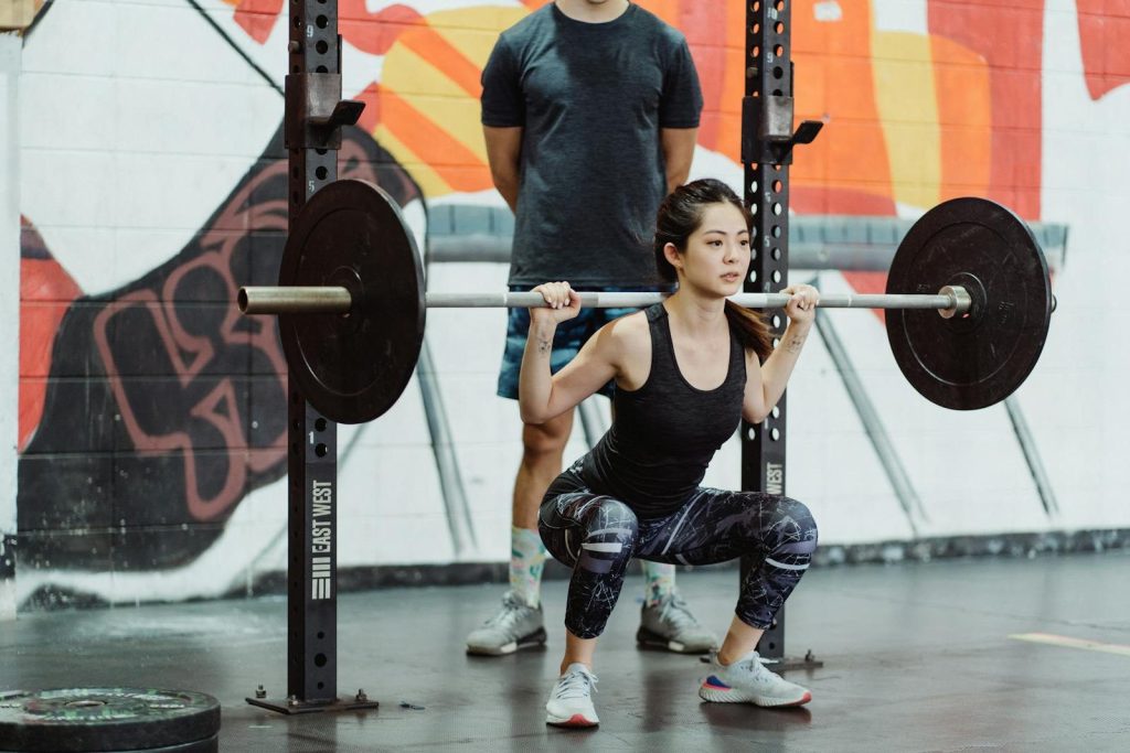 A focused young woman performs squats with weights at a gym, guided by her trainer.