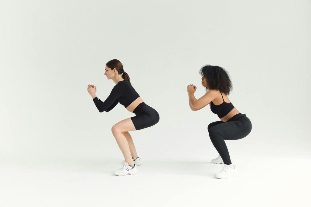 Two women performing squats in sportswear against a plain background, emphasizing fitness and a healthy lifestyle.